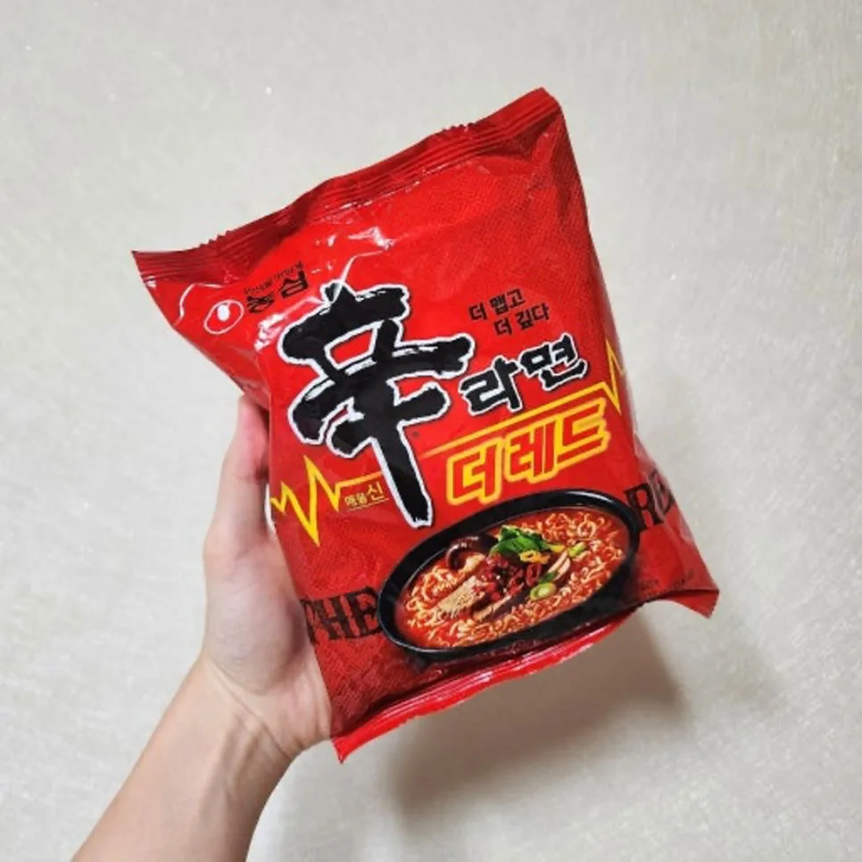 Mano che tiene confezione rossa di ramen Nongshim Shin Red con immagine di piatto di noodles