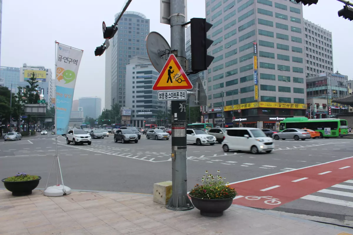 Cartello stradale a Seoul che avverte del pericolo per i pedoni che usano lo smartphone mentre attraversano la strada, con simbolo e testo in coreano.