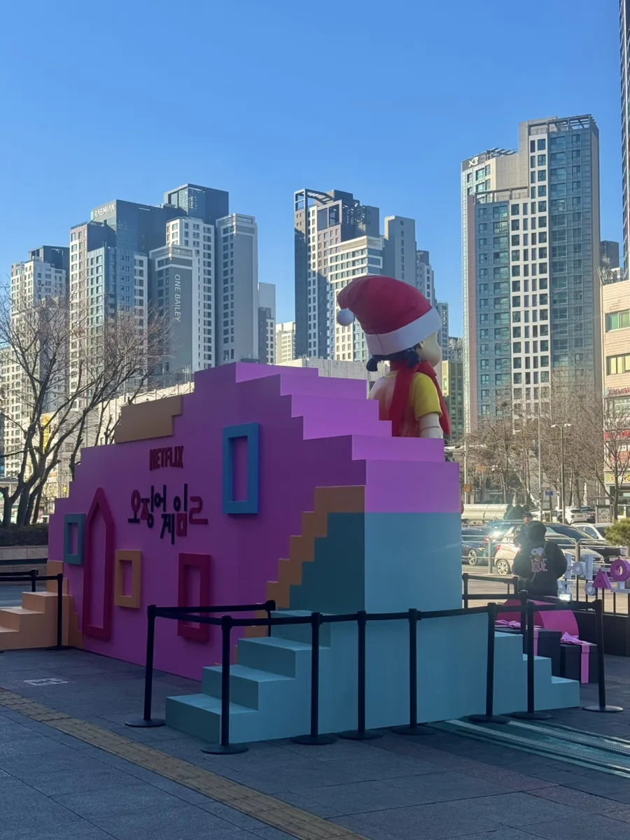 Scultura colorata ispirata a Squid Game con bambola gigante su una piattaforma in una piazza urbana