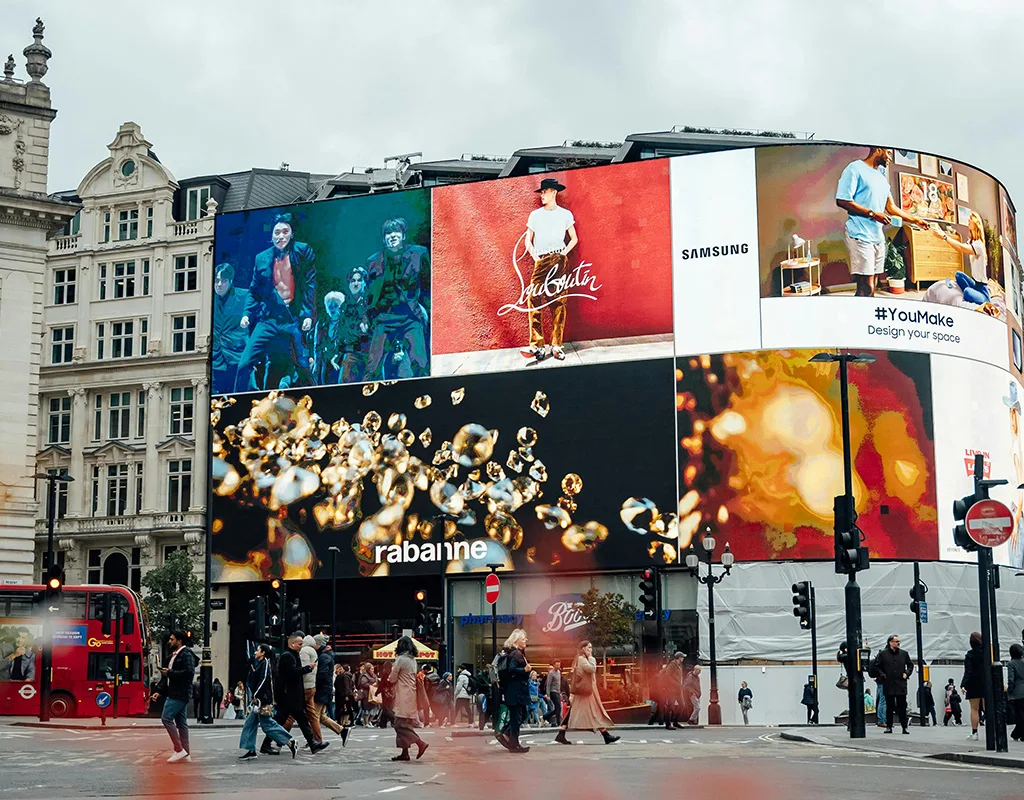 Scena diurna di Piccadilly Circus a Londra, con grandi schermi DOOH che mostrano pubblicità di vari marchi come Samsung, Rabanne e Louboutin. Persone e autobus rossi attraversano la strada affollata.