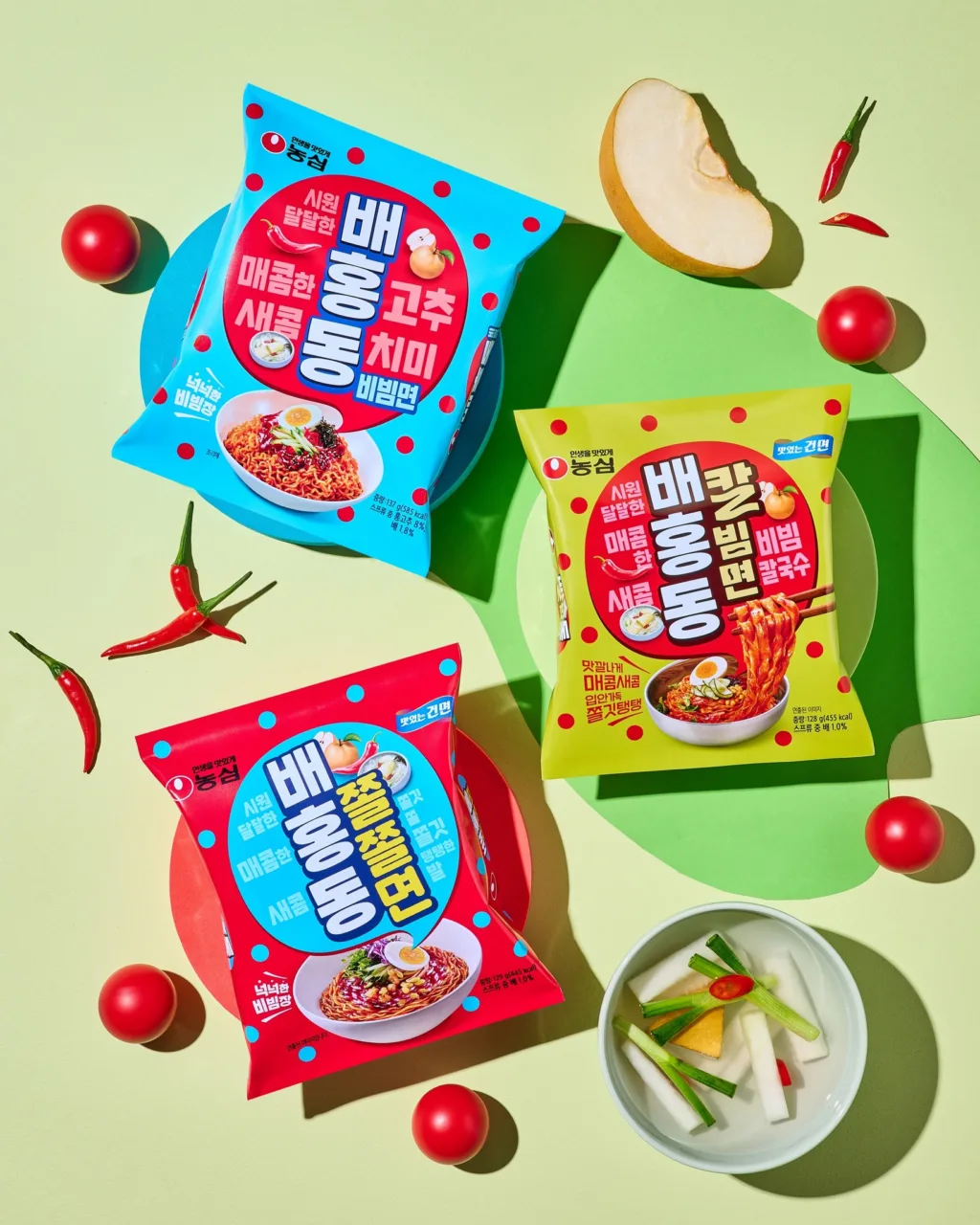 Tre confezioni di ramen coreano Nongshim Bae Hong Dong Bibimyeon in vari gusti, disposte su uno sfondo colorato con decorazioni di pomodori, peperoncini e una ciotola di kimchi bianco.
