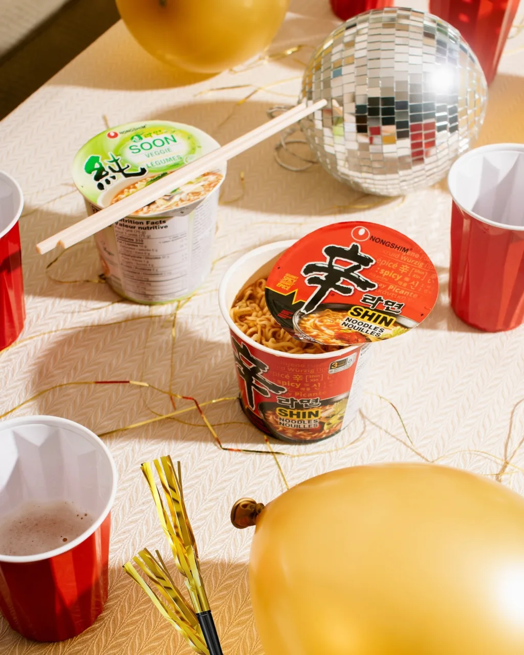 Estetica dell'Hanja nelle cup ramen Shin di Nongshim con il carattere Hanja '辛' e confezione Soon con carattere Hanja '純', circondate da decorazioni festive su una tavola chiara.