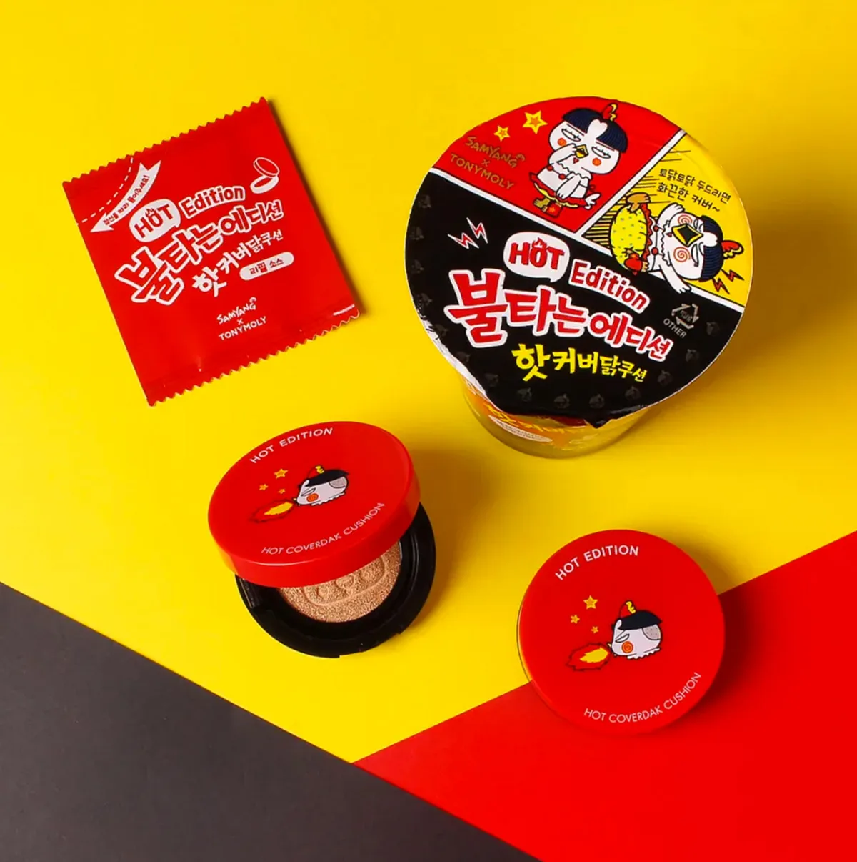 Un set di prodotti della collaborazione tra TonyMoly e Samyang, con tema "Hot Edition". L'immagine mostra un ramen istantaneo Samyang Buldak, una confezione di cushion foundation e una bustina rossa, tutti con un design ispirato al ramen piccante. Lo sfondo è giallo, rosso e nero, enfatizzando il tema del fuoco e del piccante.