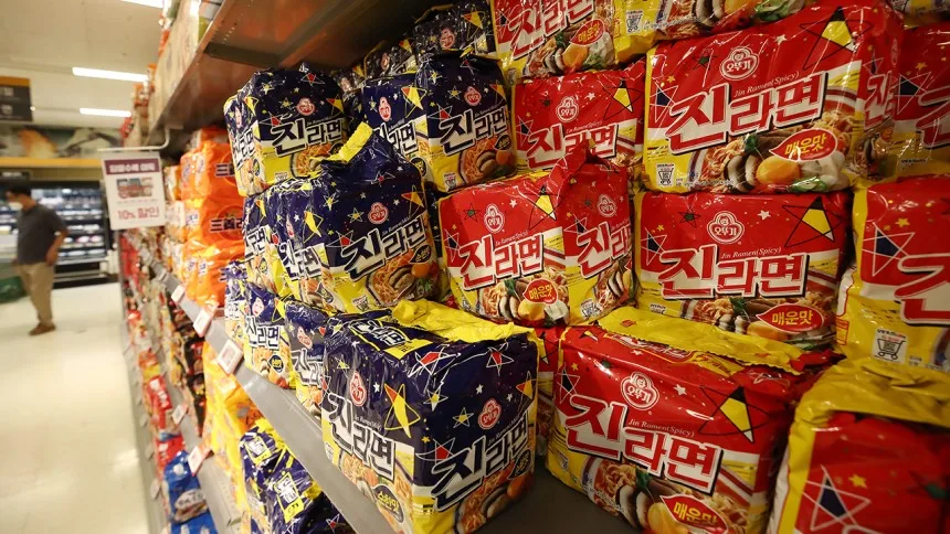 Confezioni di ramen coreano, con design colorati in blu e rosso, esposte su uno scaffale di un supermercato. Un cliente cammina sullo sfondo.