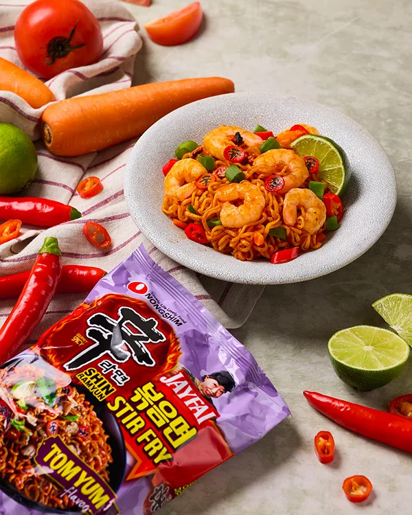 Ciotola di Shin Ramyun Stir Fry al gusto Tom Yum, servita con gamberi, peperoncino, lime e cipollotti. Accanto, la confezione del ramen su un tavolo decorato con ingredienti freschi come carote, pomodori e peperoncini rossi.
