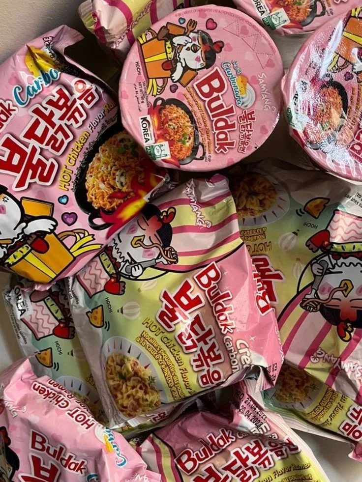Foto di diverse confezioni di ramen Buldak Hot Chicken Flavor nella variante rosa Creamy Carbonara, con il pollo mascotte "HoChi" in tema romantico.