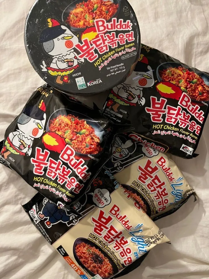 Foto di diverse confezioni di ramen Buldak Hot Chicken Flavor, inclusa la versione classica nera e la versione light beige, con il simpatico pollo mascotte "HoChi".