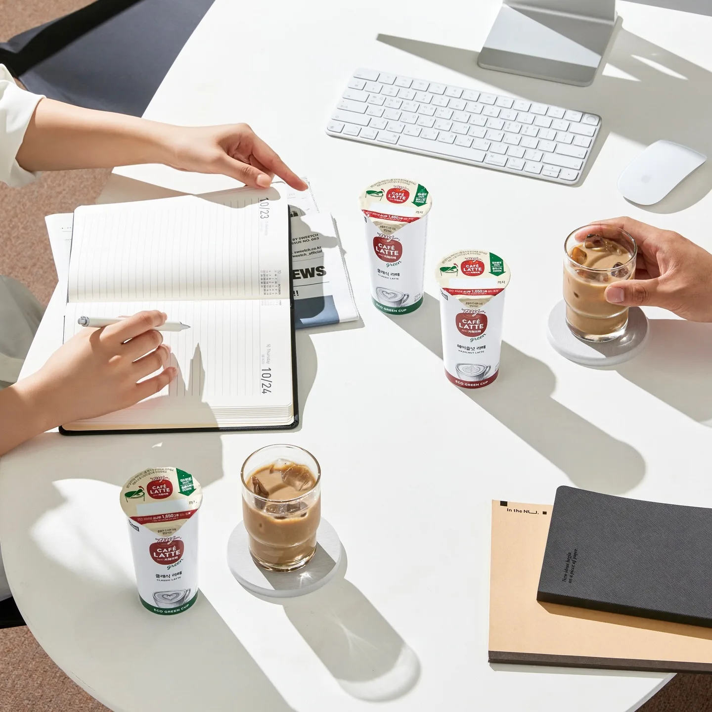Due persone sedute a un tavolo bianco da ufficio con tastiera, mouse, agenda e bicchieri di caffè freddo Maeil Café Latte; una persona scrive su un'agenda mentre l'altra tiene un bicchiere, creando un'atmosfera di lavoro e condivisione.