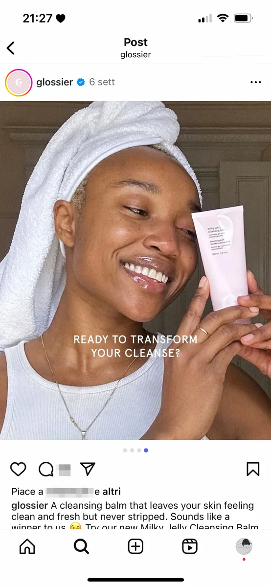 Persona sorridente con turbante bianco mostra un prodotto skincare di Glossier.