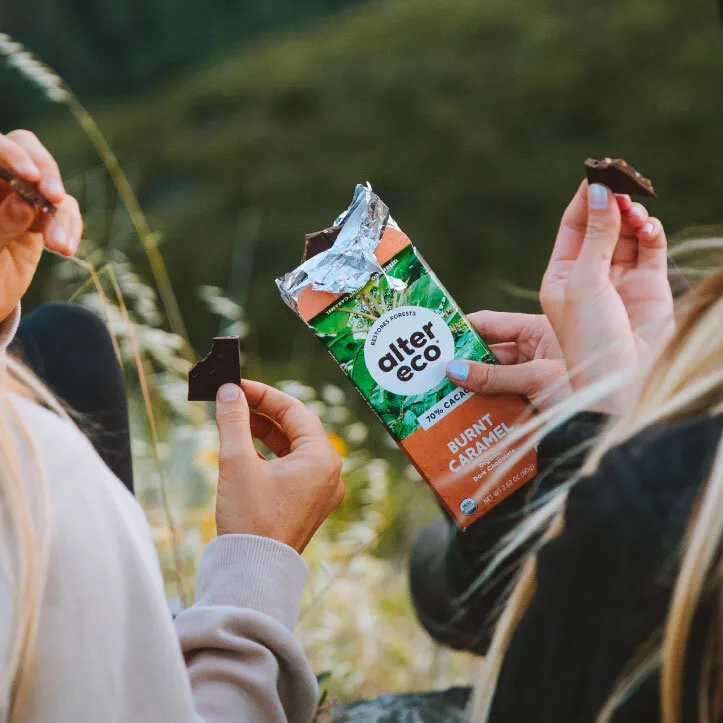 Due ragazze bionde di spalle mangiano cioccolato Alter Eco in campagna tra fiori e campi.
