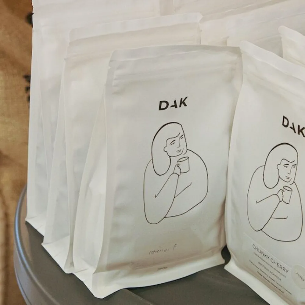 Confezioni Dak Coffee biodegradabili minimaliste con illustrazione di donna che beve caffè e logo nero su sfondo bianco.