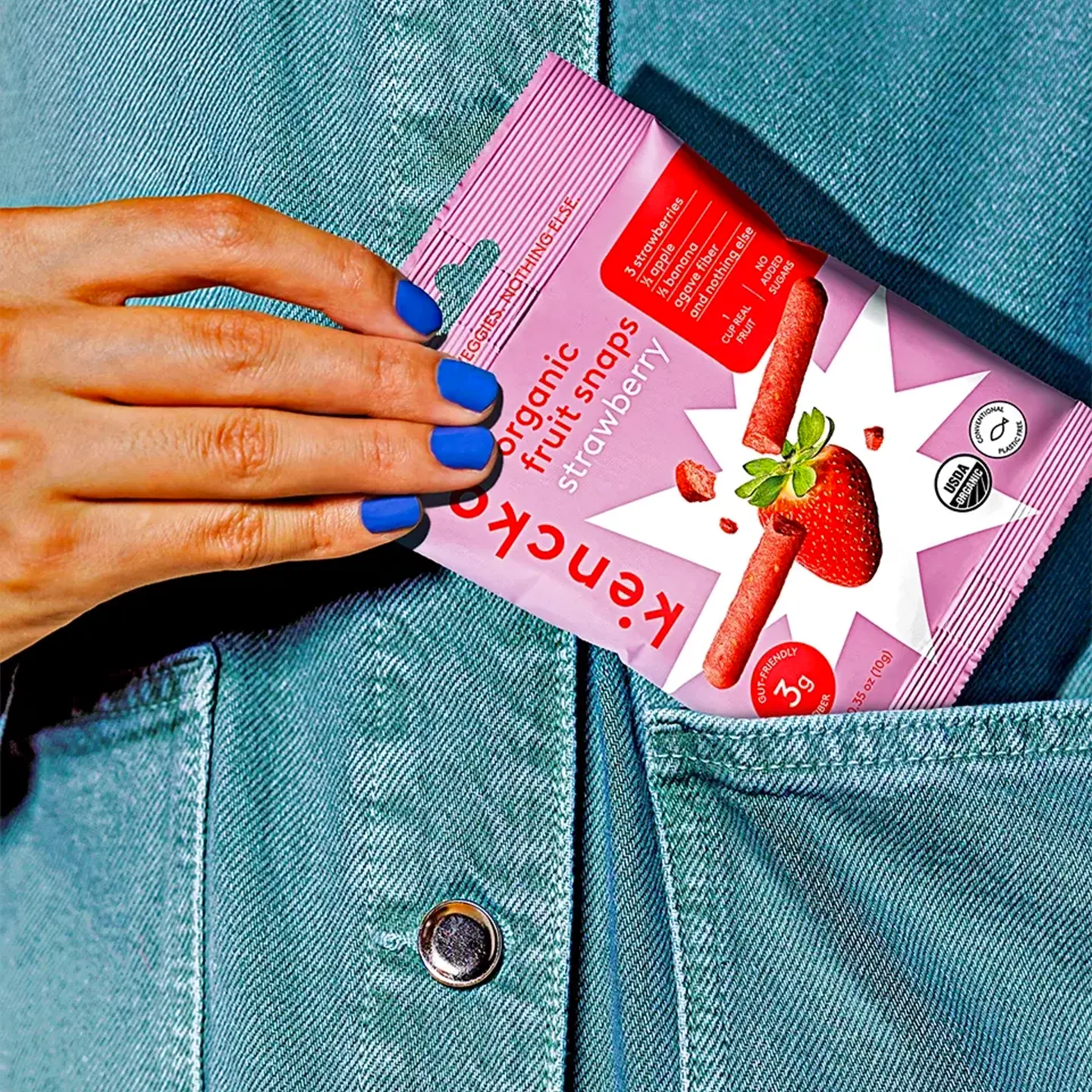 Mano con smalto blu elettrico, infila packaging Kencko rosa con fragola illustrata nel taschino di una giacca di jeans.