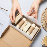 Mani che confezionano saponi artigianali in packaging sostenibile di cartone con ingredienti naturali sullo sfondo
