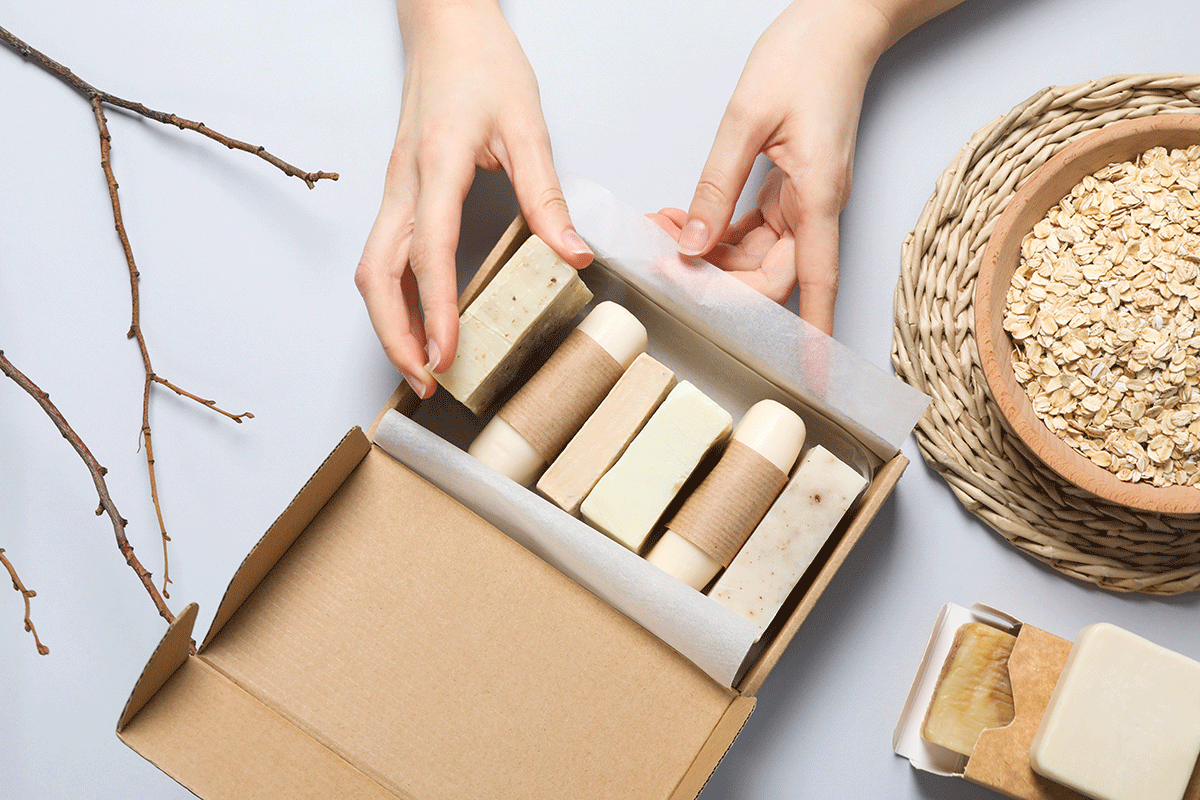Mani che confezionano saponi artigianali in packaging sostenibile di cartone con ingredienti naturali sullo sfondo