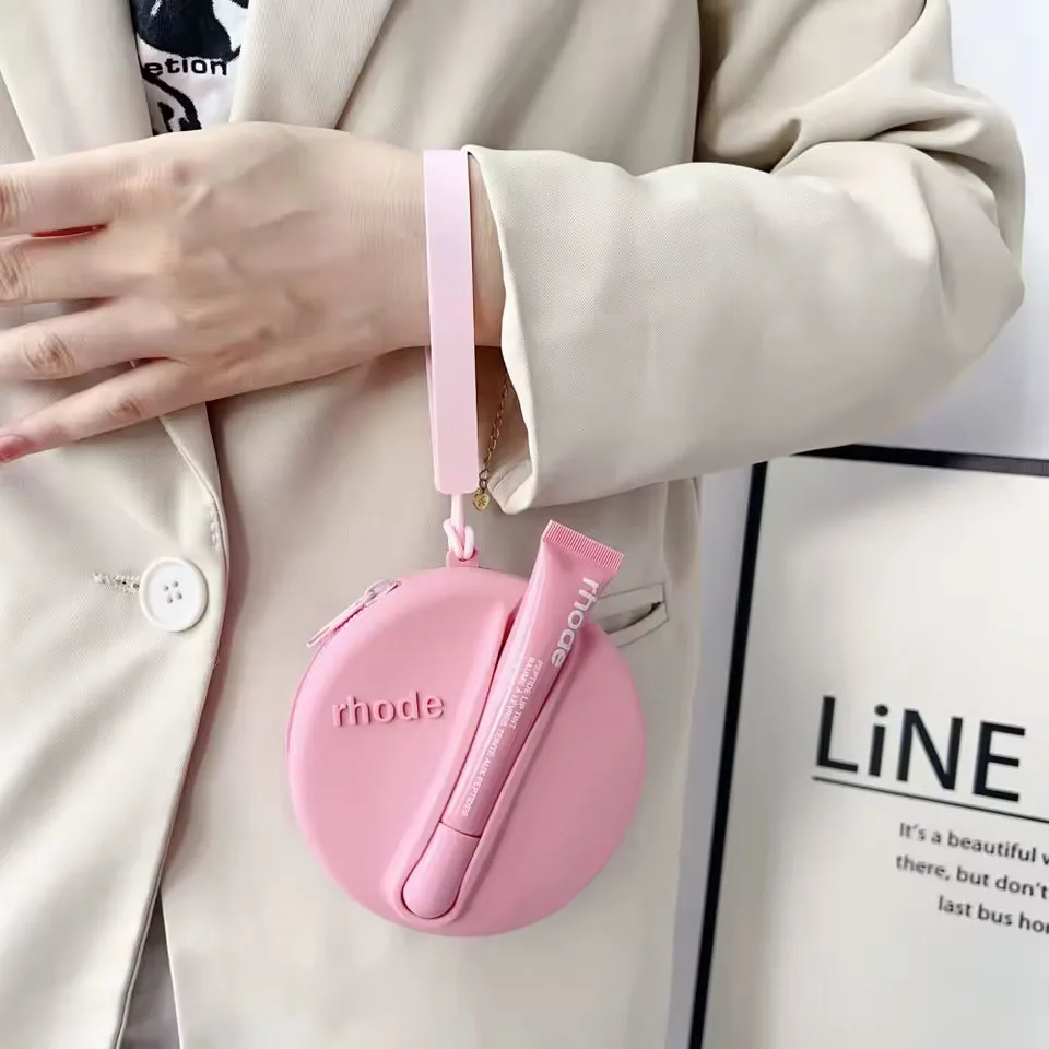Pochette tonda rosa Rhode con lipgloss applicato, indossata al polso come accessorio trendy.