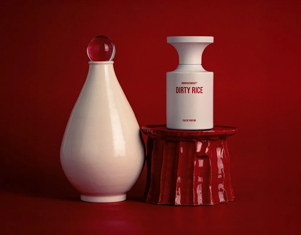 Flacone di profumo K-beauty BORNTOSTANDOUT Dirty Rice accanto a un vaso in ceramica bianca su sfondo rosso intenso