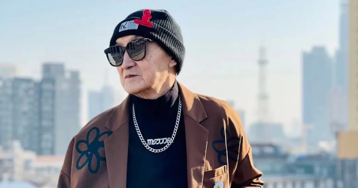 Uomo senior cinese alla moda con occhiali da sole, berretto e giacca streetwear su sfondo urbano.