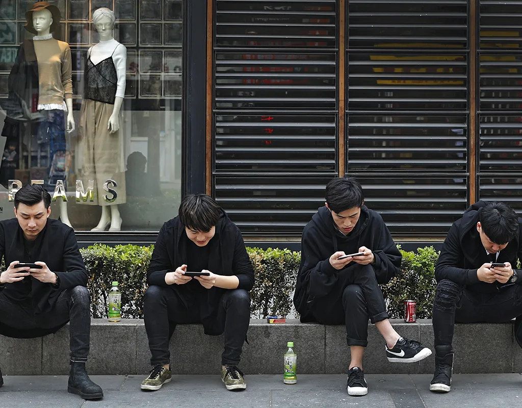 Quattro ragazzi seduti sul marciapiede davanti a una vetrina intenti a consultare i propri smartphone.