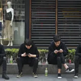 Quattro ragazzi seduti sul marciapiede davanti a una vetrina intenti a consultare i propri smartphone.