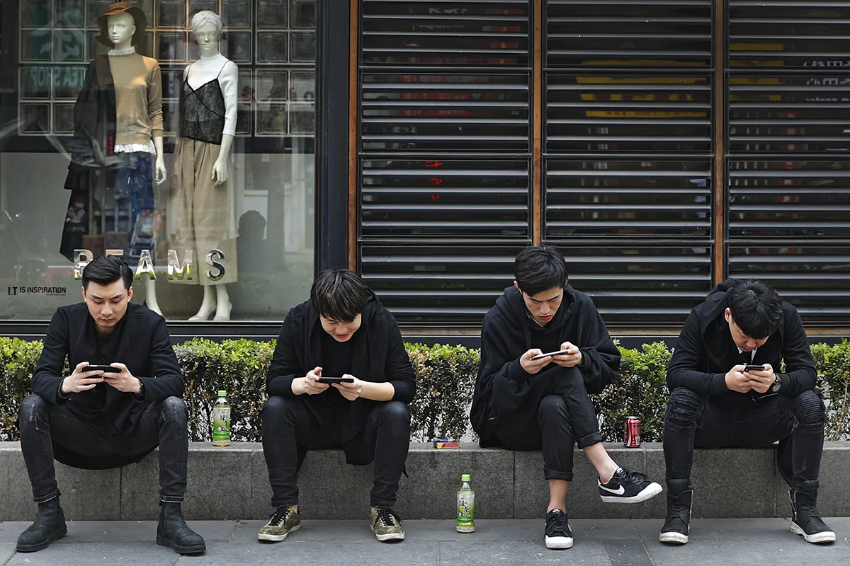 Quattro ragazzi seduti sul marciapiede davanti a una vetrina intenti a consultare i propri smartphone.