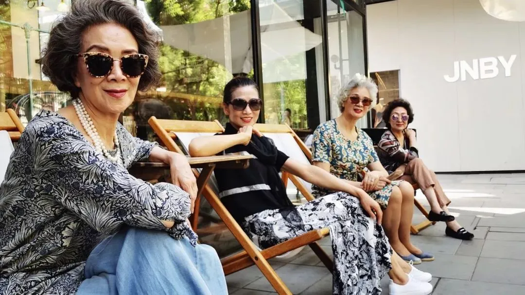 Quattro donne senior cinesi eleganti con occhiali da sole sedute su sedie a sdraio all'esterno di un negozio JNBY.